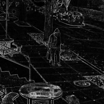 A person stands on a sidewalk near a small garden bed and a round patio table on the left, with parked cars on the street in front of a house at night.