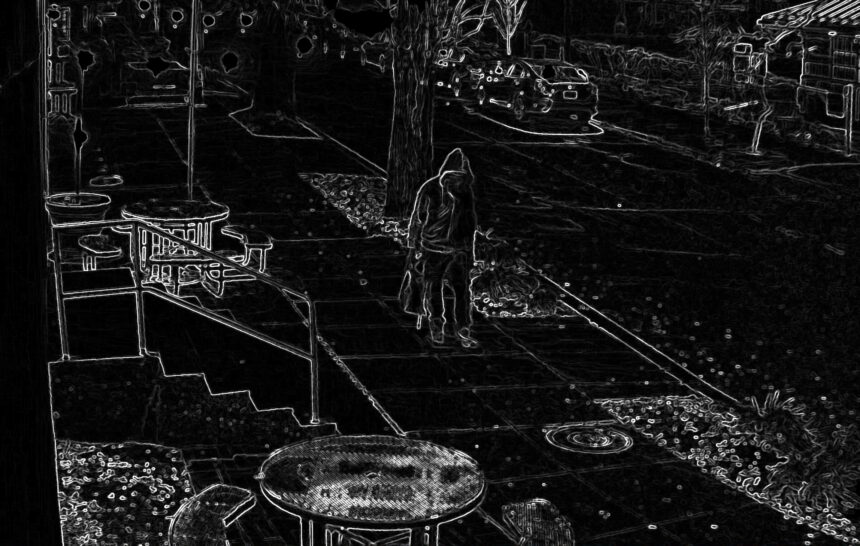 A person stands on a sidewalk near a small garden bed and a round patio table on the left, with parked cars on the street in front of a house at night.