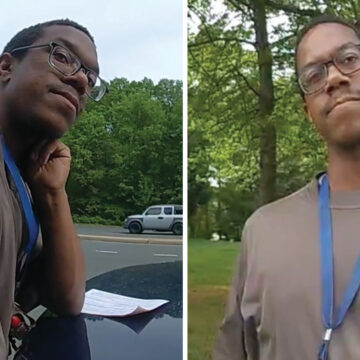 Man with glasses and a blue lanyard outdoors, leaning on a car hood and holding papers near a parking lot trees in the background.